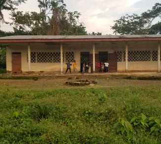 Ein einstöckiges Schulgebäude in Beoko mit einer langen Veranda, mehreren Türen und Fenstern mit Gittermustern. Vor der Schule stehen einige Kinder in Gruppen beisammen. Die Umgebung ist grün mit Gras und Büschen im Vordergrund, während Bäume im Hintergrund wachsen. Dieses Schulprojekt wird von der Oldtimer Rallye Oberschwaben Classic unterstützt.