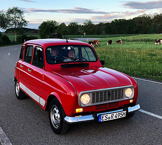 Ein roter Renault 4 mit deutschem Oldtimer-Kennzeichen fährt auf einer kurvigen Landstraße durch eine idyllische Landschaft Oberschwabens. Zwei fröhliche Personen sitzen im Wagen und genießen die Fahrt. Rechts neben der Straße grasen Kühe auf einer grünen Wiese. Der Himmel ist blau mit wenigen Wolken, und die Abendstimmung sorgt für eine warme, nostalgische Atmosphäre.